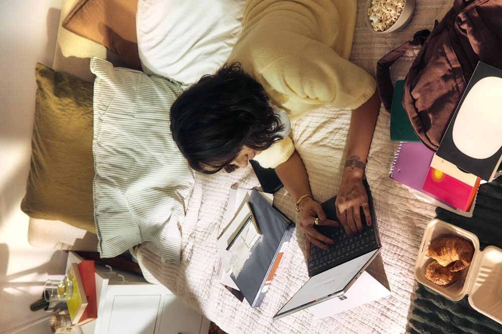 Woman working on laptop on bed with snacks and books